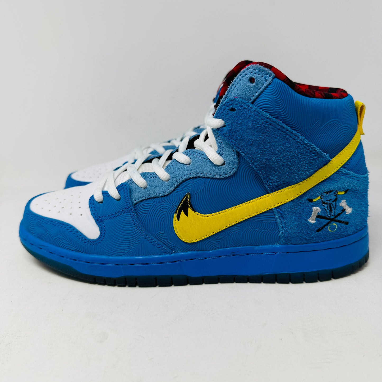 The Nike SB Dunk High Familia Blue Ox, a 2015 release by Nike, features clean blue uppers with a yellow swoosh, decorative stitching, red and black checkered lining, lace-up front, and an edgy skull with crossed bones on the side—its standout design loses no stars.