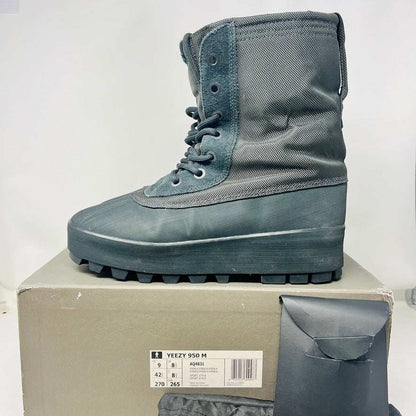 A 2015 Adidas Yeezy 950 Pirate Black boot is displayed on its box against a plain white background. This authentic Yeezy features a high lace-up design and sturdy ridged soles. The box includes size details, a partially visible label, and extra laces for added versatility.