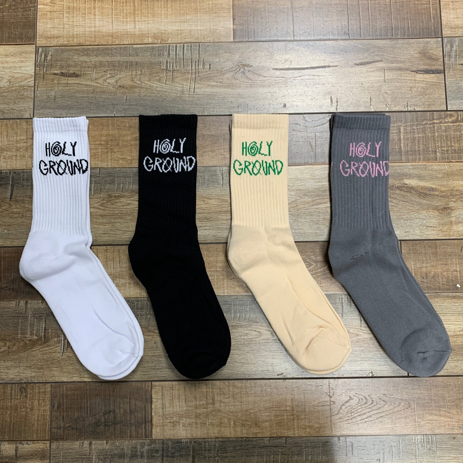 Holy Ground Socks in white, black, beige, and gray displayed on a wooden floor.