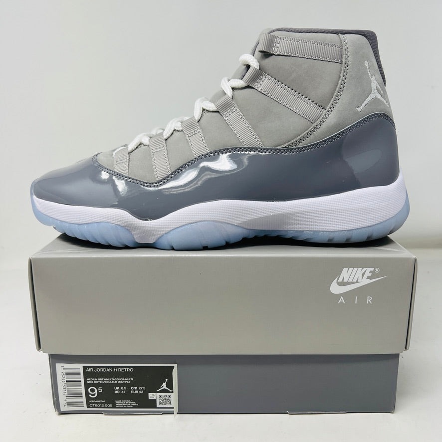 A pair of Jordan 11 Retro Cool Grey (2021) sneakers with white laces and translucent soles rest on a gray Nike Air box, showcasing the shiny patent leather and Jumpman logo. Size 9.5 appears on the label.