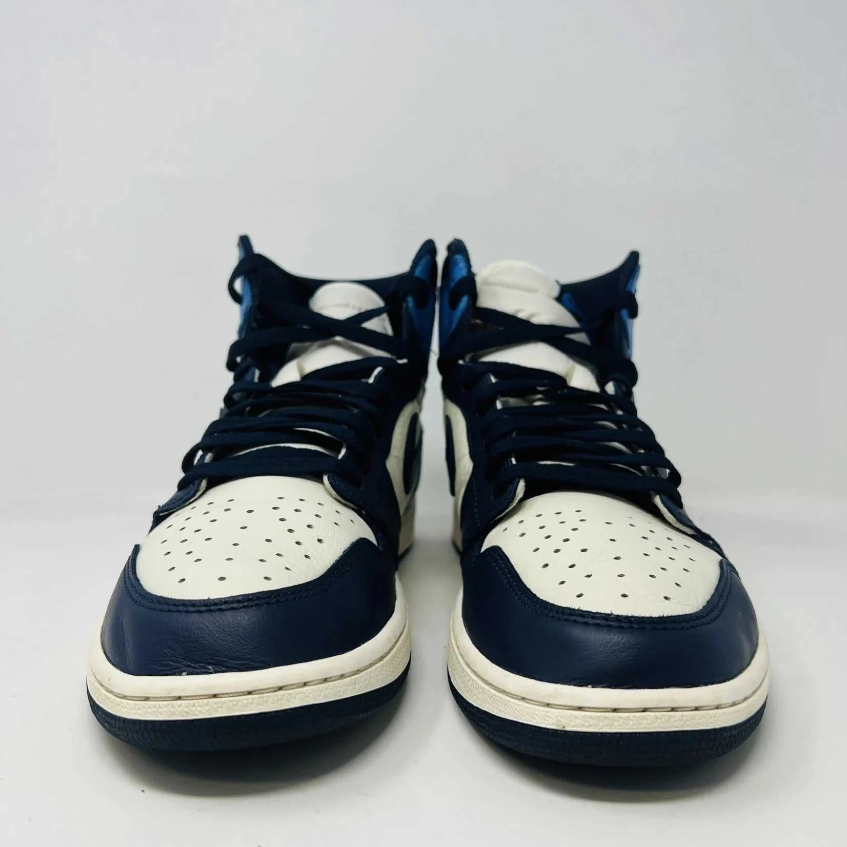 The Jordan 1 Retro High Obsidian sneakers, featuring a blue, white, and black design with signature swoosh and Air Jordan branding on the ankle, are gracefully displayed on a black Nike box with a red logo. A bag of blue shoelaces accompanies the 2019 release from Jordan.