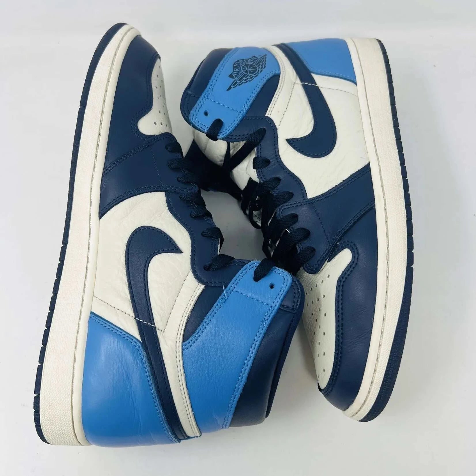 The Jordan 1 Retro High Obsidian sneakers, featuring a blue, white, and black design with signature swoosh and Air Jordan branding on the ankle, are gracefully displayed on a black Nike box with a red logo. A bag of blue shoelaces accompanies the 2019 release from Jordan.