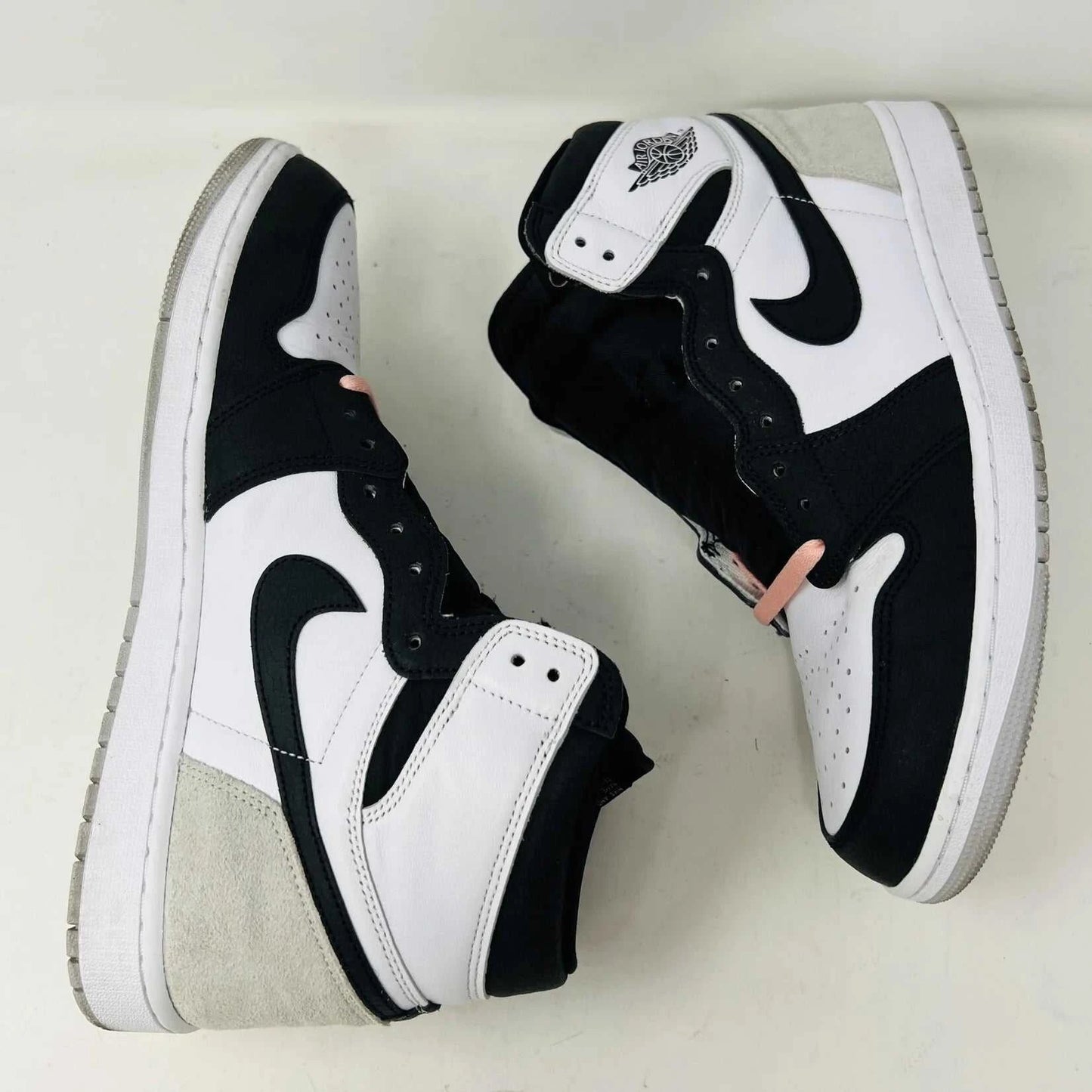 The Jordan 1 Retro High OG Bleached Coral sneaker by Jordan features a high-top design with a clean white base, prominent black Nike swoosh, gray detailing on the heel, and the iconic Jordan wings logo near the ankle. Its set against a plain background.