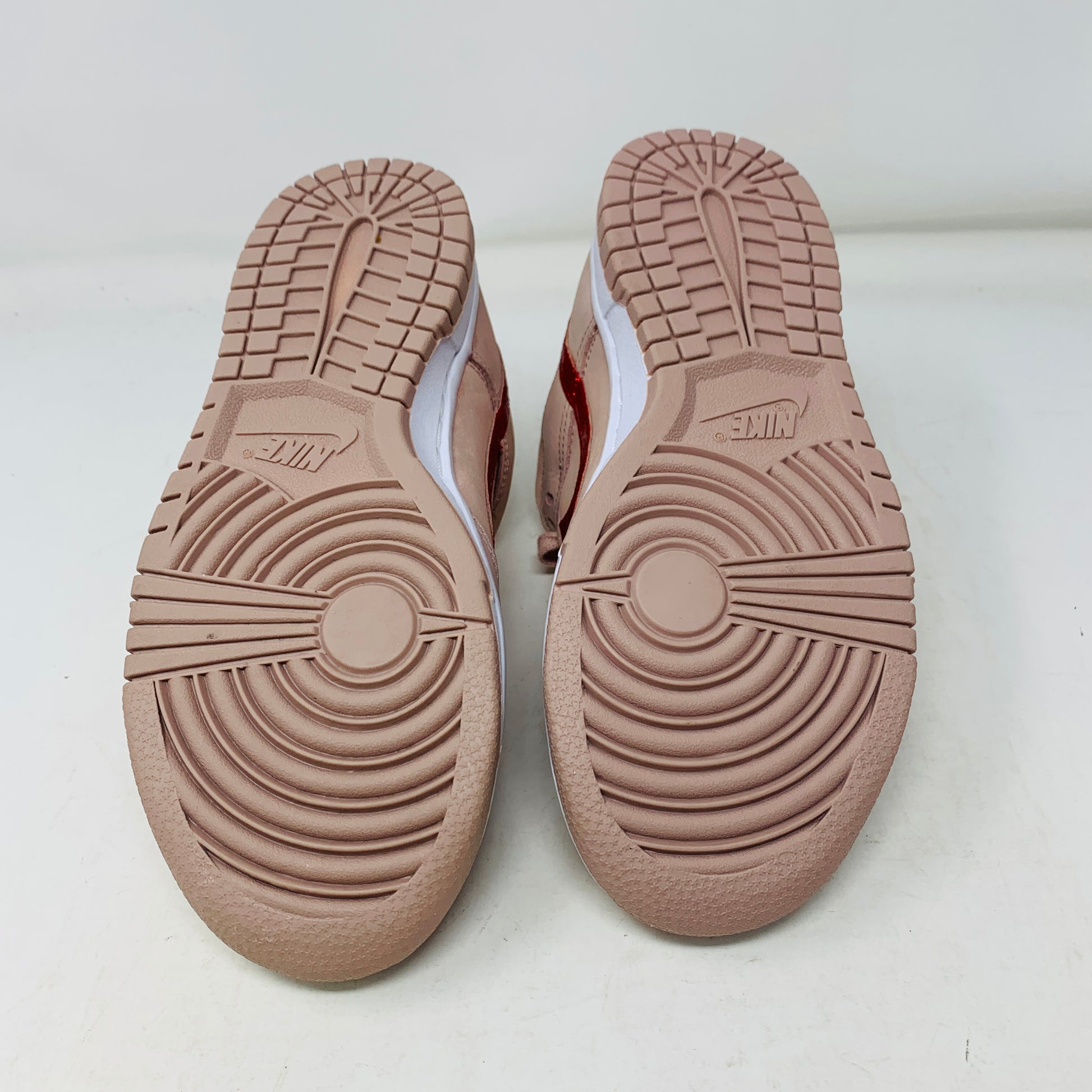 The image displays the Nike Dunk Low PRM Pink Oxford (W) sneakers, featuring a pinkish-brown tread pattern on a white background. The circular and linear designs enhance grip, complete with the Nike logo on each sole.