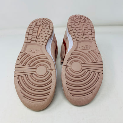 The image displays the Nike Dunk Low PRM Pink Oxford (W) sneakers, featuring a pinkish-brown tread pattern on a white background. The circular and linear designs enhance grip, complete with the Nike logo on each sole.