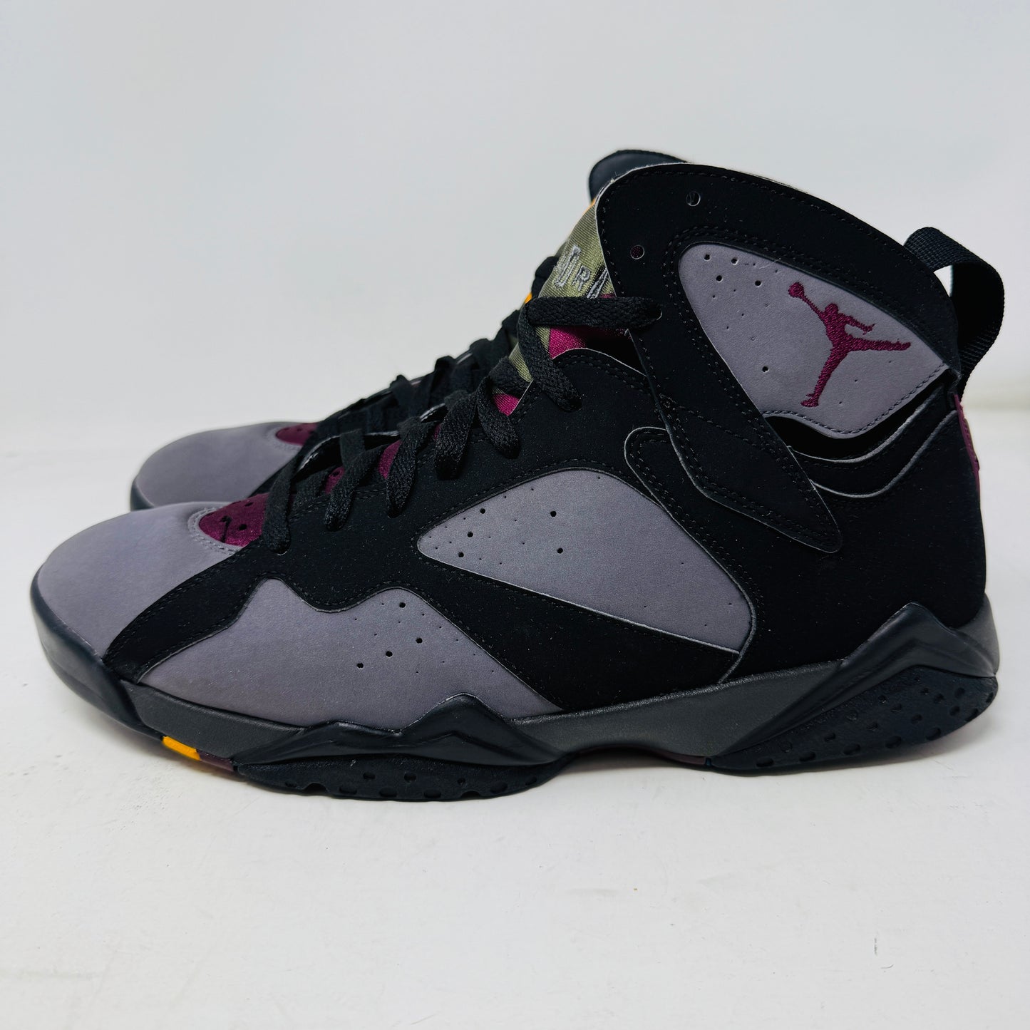 The Jordan 7 Retro Bordeaux (2015) sneakers, in black and gray with maroon accents and the Jumpman logo, feature angular midsoles, dark laces, and sleek uppers. They evoke a 2015 vibe and are photographed side by side on a white background.