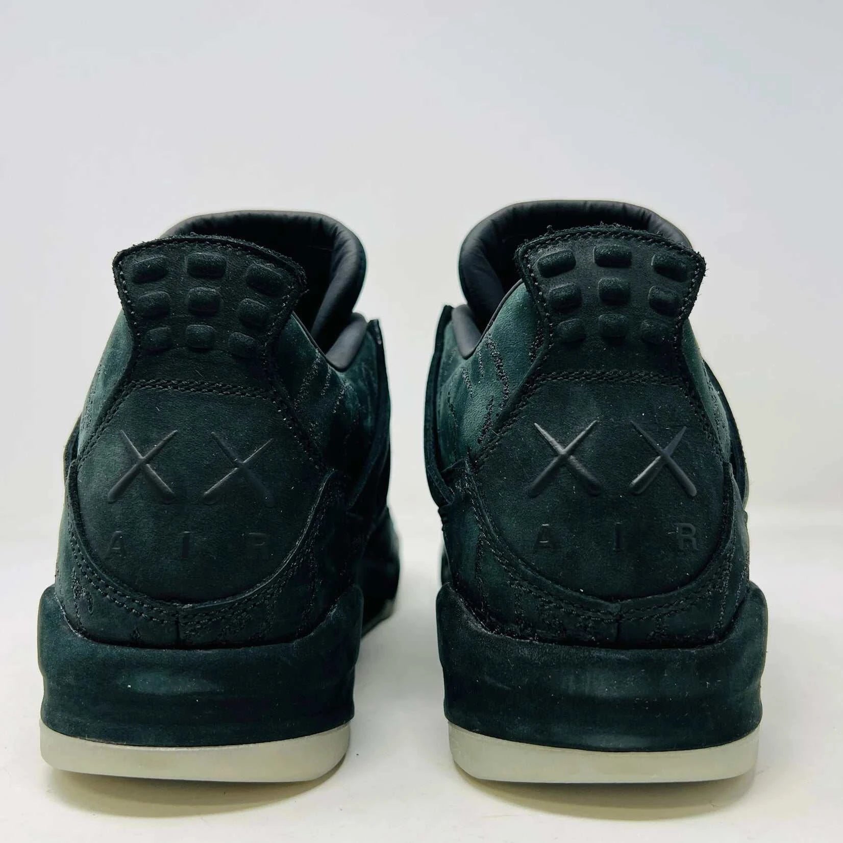 The Jordan 4 Kaws Black sneaker, brand new with distinct stitching and a sculpted midsole, sits atop its branded shoe box. Complete with textured suede details and accompanied by a dustbag, it rests on a black cloth beneath.