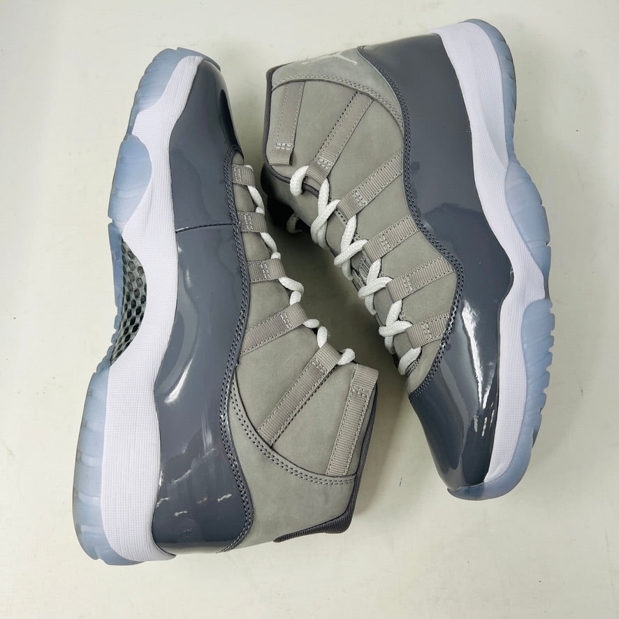 A pair of Jordan 11 Retro Cool Grey (2021) sneakers with white laces and translucent soles rest on a gray Nike Air box, showcasing the shiny patent leather and Jumpman logo. Size 9.5 appears on the label.