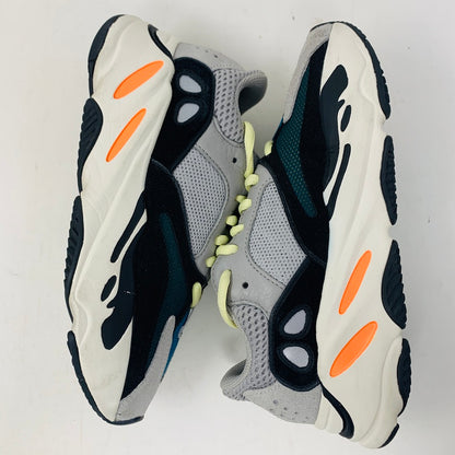 A pair of adidas Yeezy Boost 700 Wave Runner sneakers in gray and black, with white soles and orange accents, are displayed side by side on a white surface, highlighting their unique design and prominent midsoles.