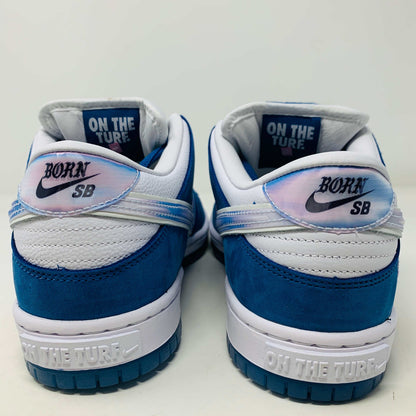 The Nike SB Dunk Low Born X Raised One Block At A Time sneaker, in blue and white with a metallic swoosh and embroidered heel details, sits on a Nike shoebox. It includes three pairs of shoelaces—two white and one pink—in front of the 2023 edition box.