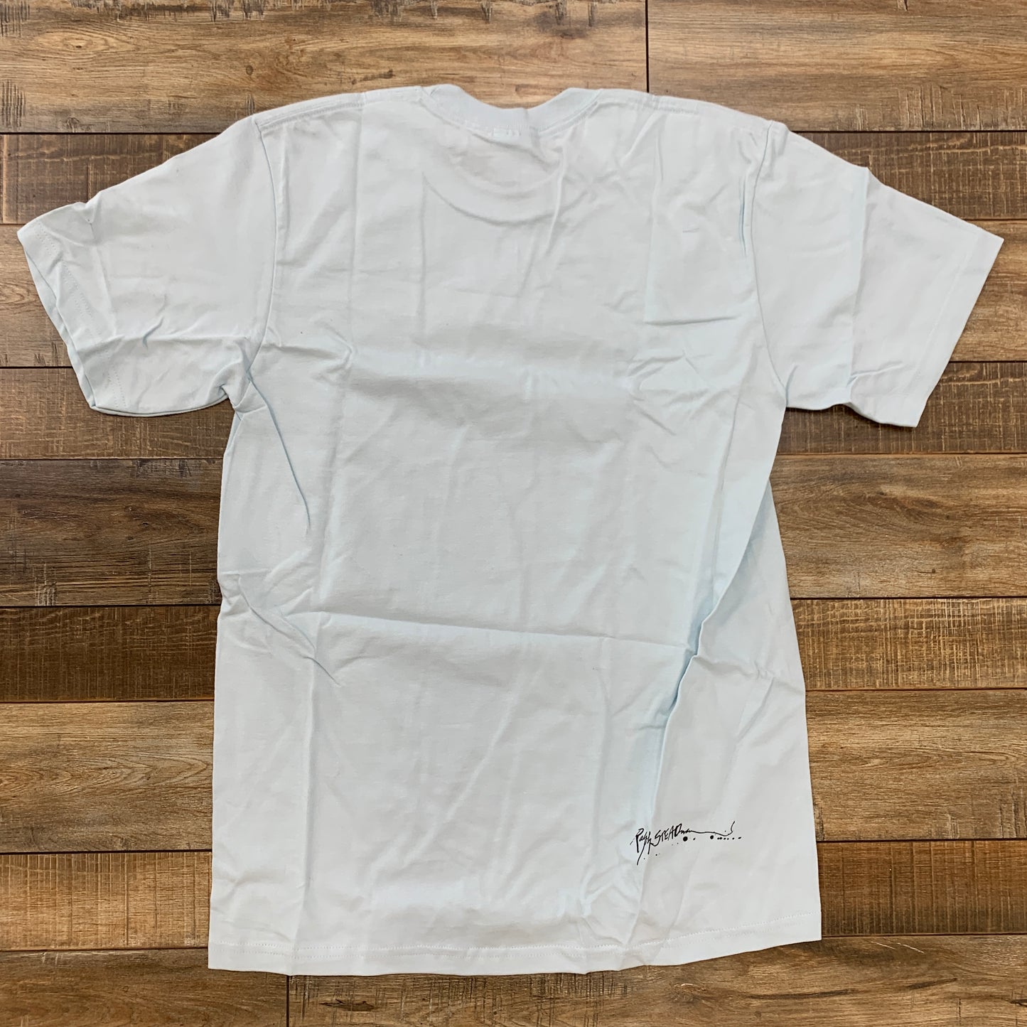 A Pale Blue Supreme Ralph Steadman Box Logo Tee is laid out flat on a wooden floor, with a distinct small signature or design near the bottom, but otherwise remains plain.