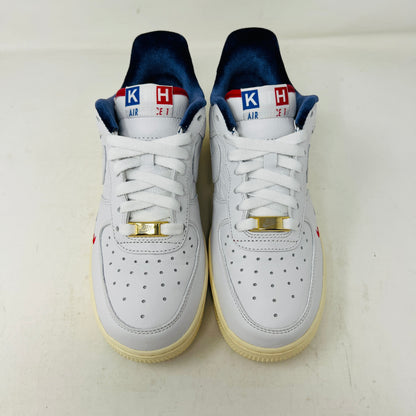 The Nike Air Force 1 Low Kith Paris sneakers are white with perforated toe boxes, a blue interior, red and white K and H tongue patches, gold lace accessories, and slightly yellowed soles.