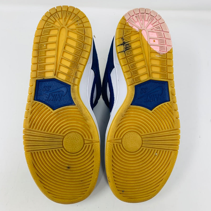 The image features the soles of Nike SB Dunk Low Los Angeles Dodgers sneakers. The soles combine yellow and pink rubber with distinct grip patterns, displaying the blue Nike logo on each shoe. The background is plain white.