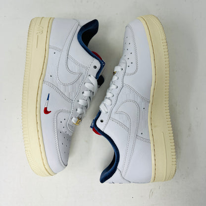 The Nike Air Force 1 Low Kith Paris sneakers have white exteriors, cream-colored soles, and blue inner lining. They feature a red and blue Nike swoosh on the side, placed side by side on a plain white background.