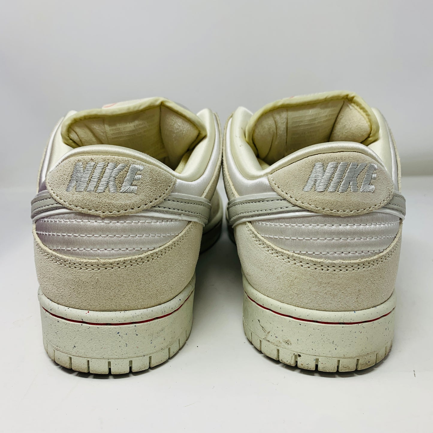 The image showcases a pair of Nike SB Dunk Low City of Love Light Bone sneakers from the back, with the Nike logo embroidered in gray on the heel. The white shoes feature a classic design, textured soles, and show signs of slight wear.