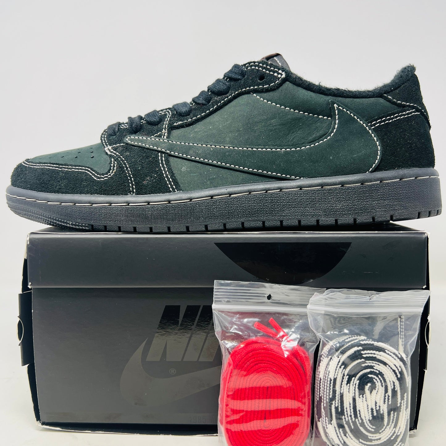 The Jordan 1 Retro Low OG SP Travis Scott Black Phantom sneakers feature clean black uppers with a large Swoosh, come in a matching black box, and include two extra lace sets—red and black-and-white—packaged in plastic bags beside the box.