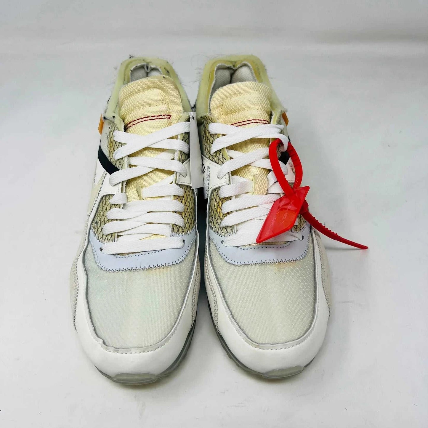 The Nike Air Max 90 Off-White sneakers, featuring a prominent red zip tie, translucent soles, and distinct stitched details with white and beige mesh accents, are displayed on a plain background.