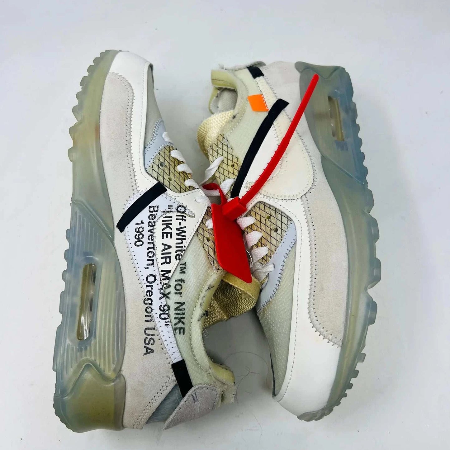 The Nike Air Max 90 Off-White sneakers, featuring a prominent red zip tie, translucent soles, and distinct stitched details with white and beige mesh accents, are displayed on a plain background.