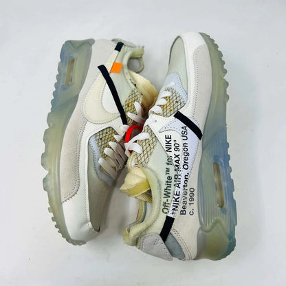 The Nike Air Max 90 Off-White sneakers, featuring a prominent red zip tie, translucent soles, and distinct stitched details with white and beige mesh accents, are displayed on a plain background.