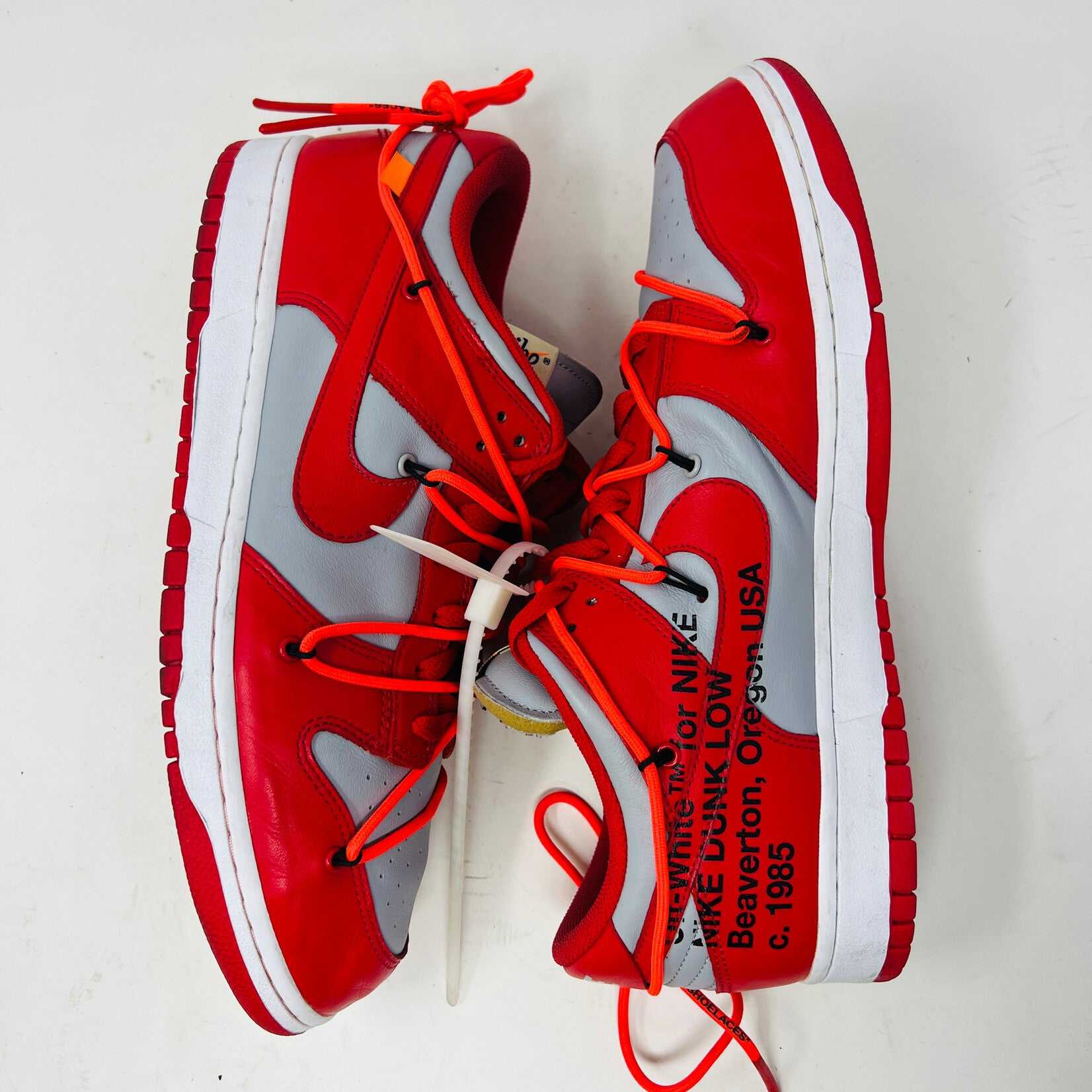 A red and gray Nike Dunk Low Off-White University Red sneaker with orange laces sits atop a shiny silver box. These authentic shoes feature clean uppers, a red swoosh, overlapping lace loops, visible tags, and a unique design.