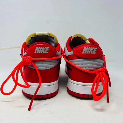 A red and gray Nike Dunk Low Off-White University Red sneaker with orange laces sits atop a shiny silver box. These authentic shoes feature clean uppers, a red swoosh, overlapping lace loops, visible tags, and a unique design.