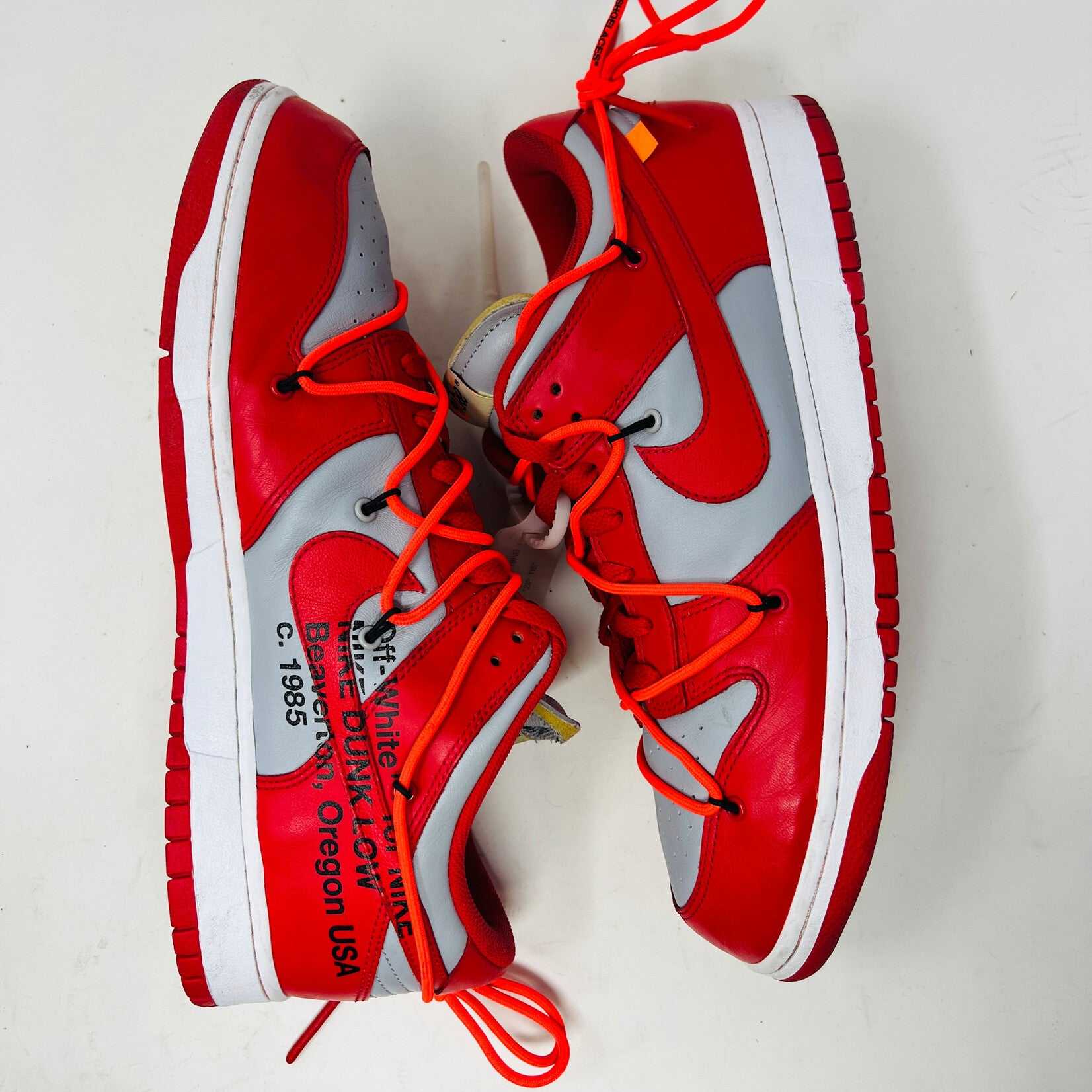 A red and gray Nike Dunk Low Off-White University Red sneaker with orange laces sits atop a shiny silver box. These authentic shoes feature clean uppers, a red swoosh, overlapping lace loops, visible tags, and a unique design.