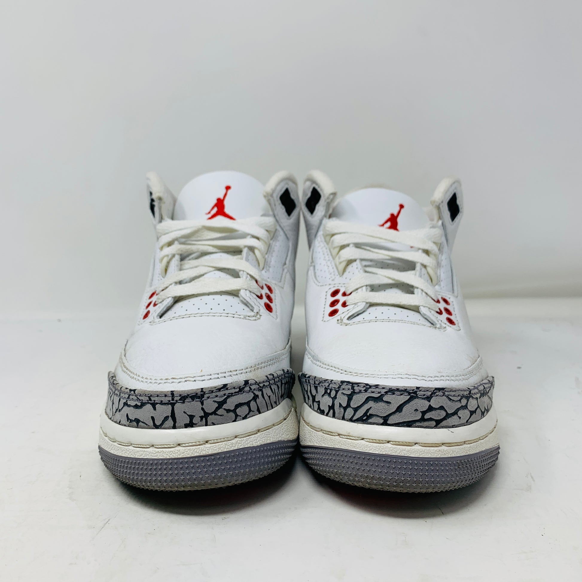 The Jordan 3 Retro White Cement Reimagined (GS) by Jordan are white and gray sneakers with red accents, showcasing an elephant print on the front and a visible logo on the tongue, set against a plain white background.