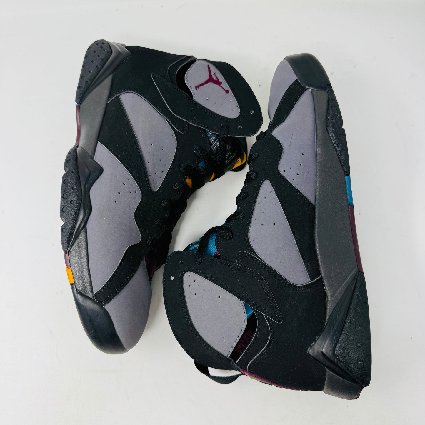 The Jordan 7 Retro Bordeaux (2015) sneakers, in black and gray with maroon accents and the Jumpman logo, feature angular midsoles, dark laces, and sleek uppers. They evoke a 2015 vibe and are photographed side by side on a white background.