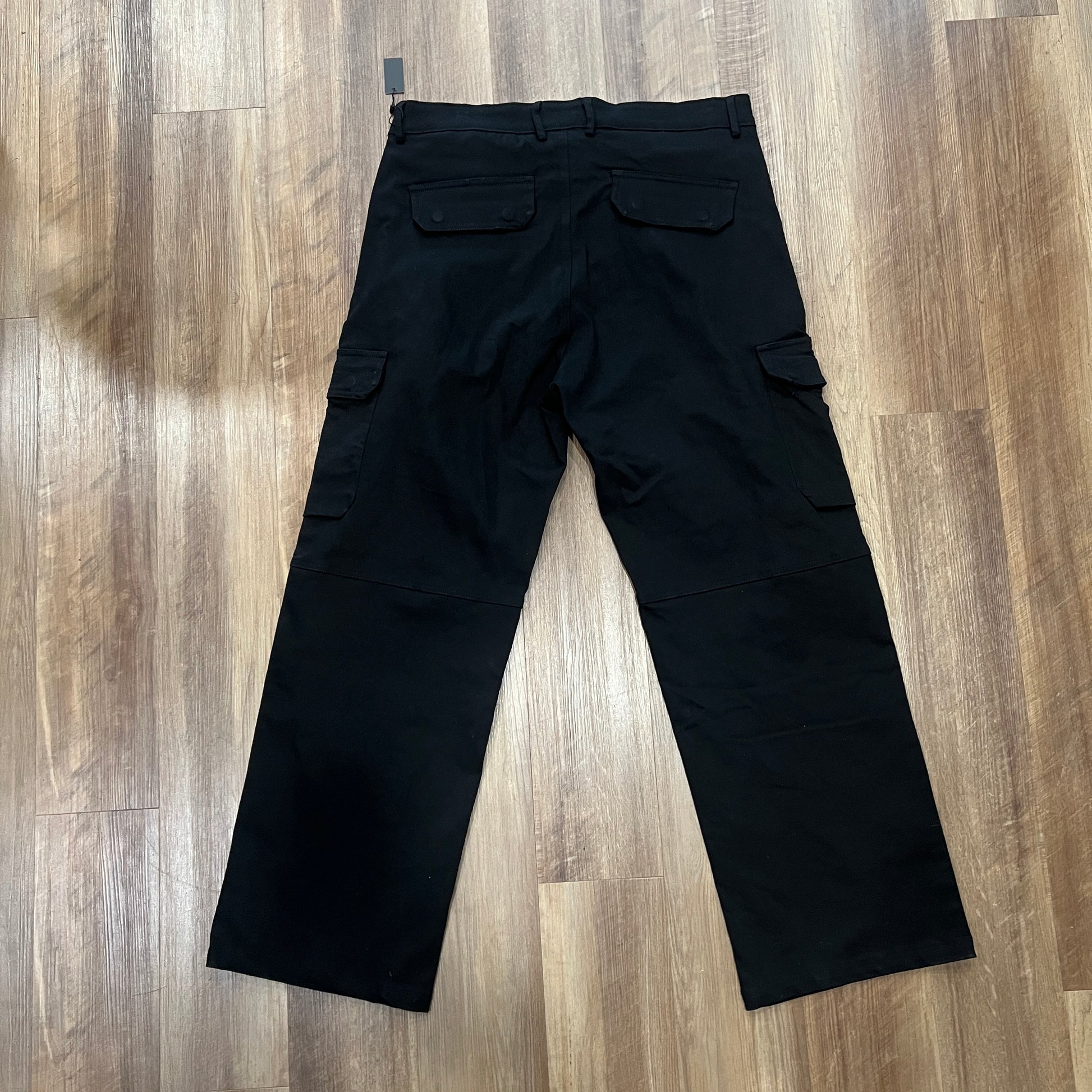 The Holy Ground Cargo Black pants, featuring large side pockets and a subtle shine, are on a wooden floor. Available in XS-XXL with a white logo on one pocket and a visible waist tag. Be sure to consult the size chart for the perfect fit.