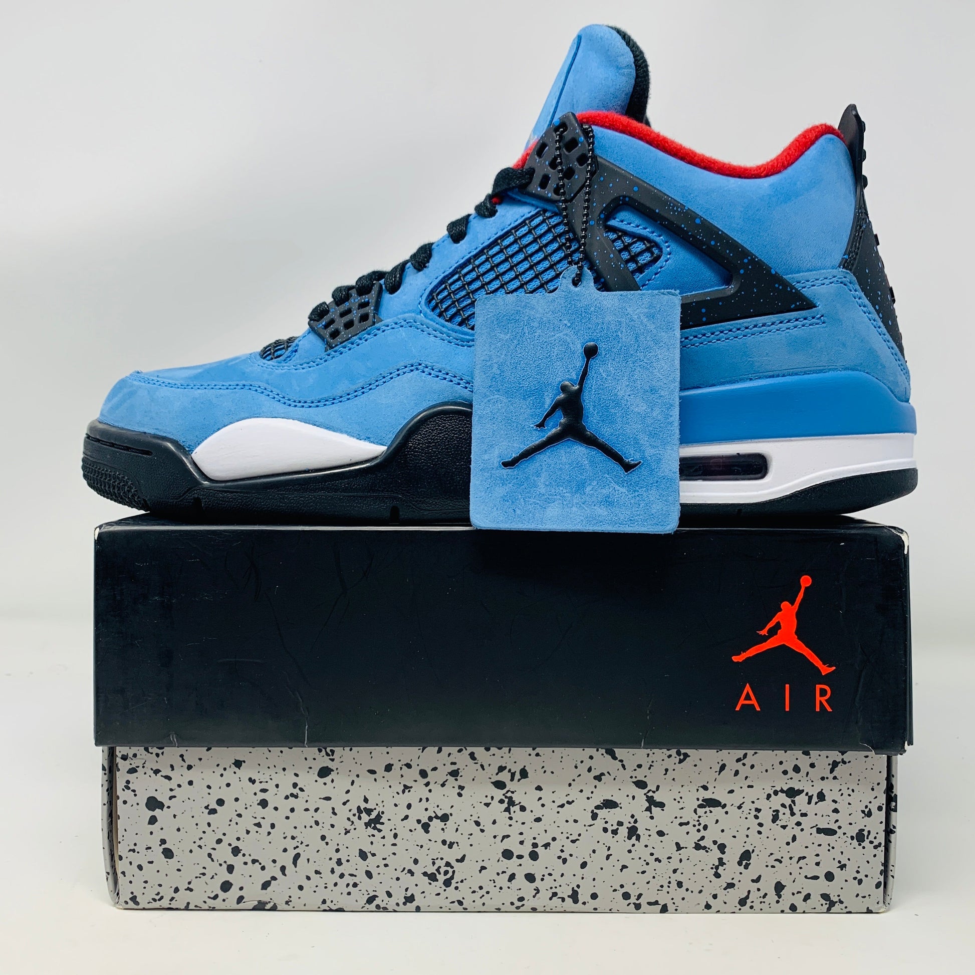 The Jordan 4 Retro Travis Scott Cactus Jack sneaker in blue, accented with black and red, features a visible air unit in the heel and a Jumpman logo hang tag, packaged in a black speckled box with an orange Jumpman logo and AIR.