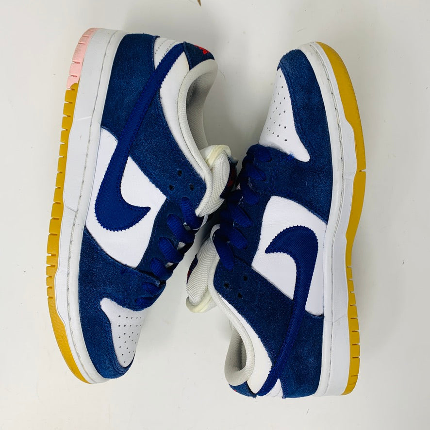 The Nike SB Dunk Low Los Angeles Dodgers sneakers, featuring blue suede and white leather panels with white laces and yellow soles, have a distinctive blue swoosh and a contrasting pink heel section. They are displayed side by side on a white background.