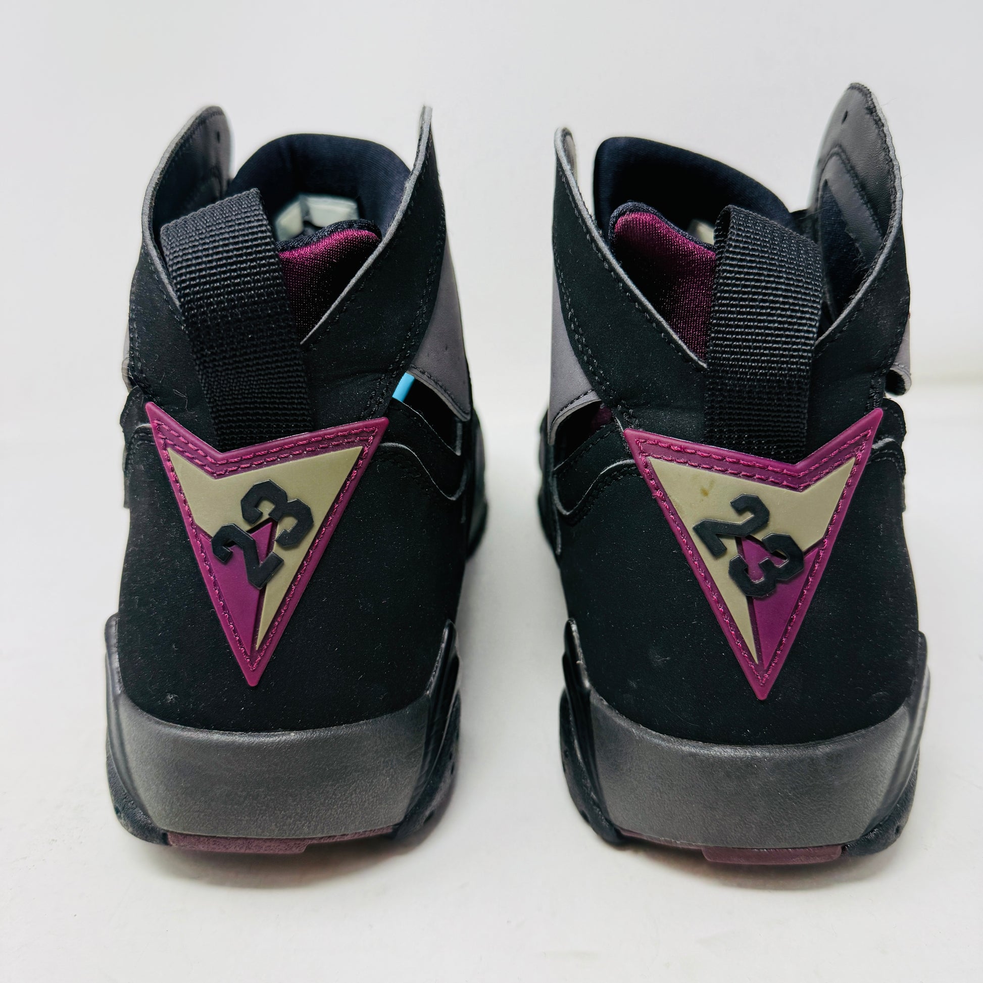 The Jordan 7 Retro Bordeaux (2015) sneakers, in black and gray with maroon accents and the Jumpman logo, feature angular midsoles, dark laces, and sleek uppers. They evoke a 2015 vibe and are photographed side by side on a white background.