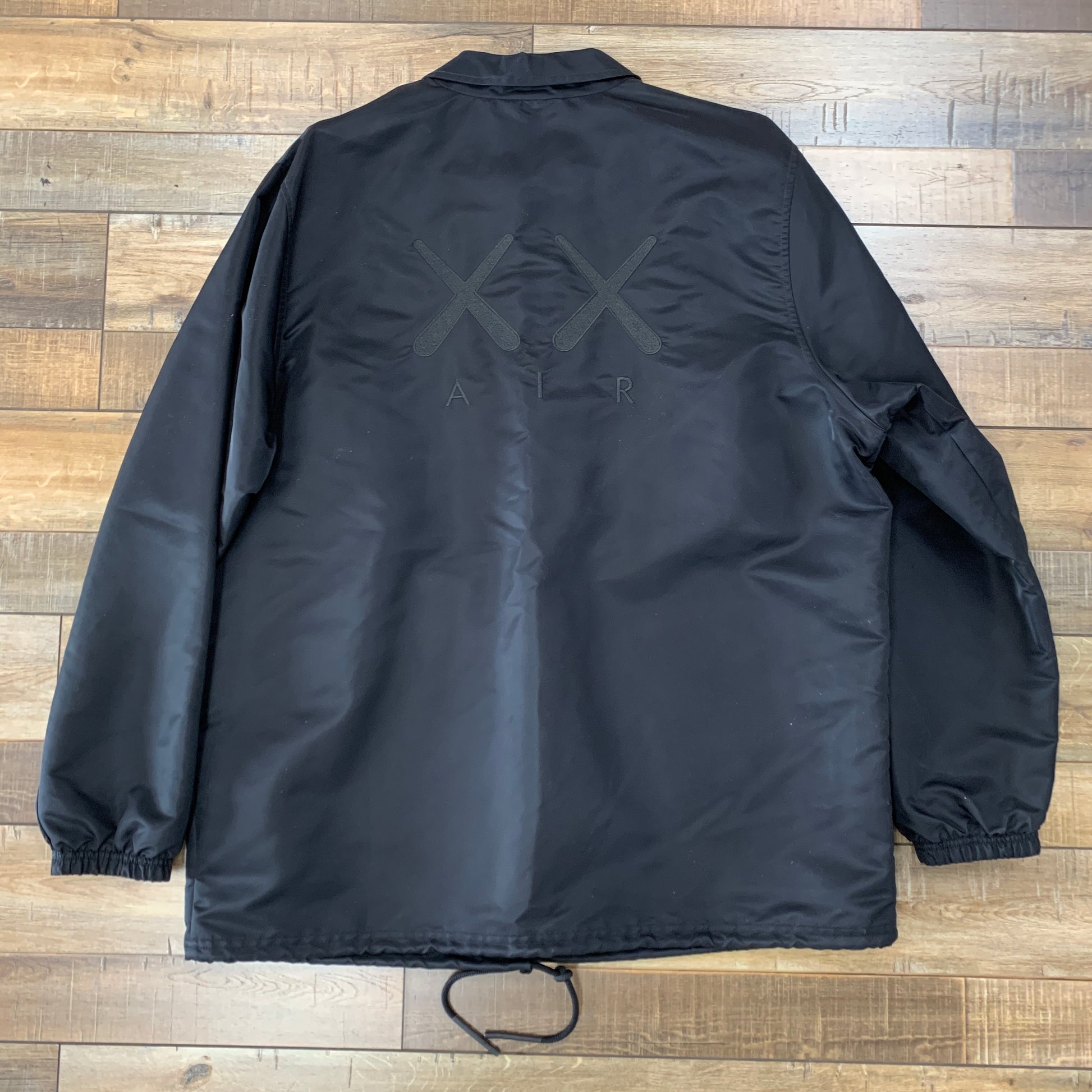 The KAWS x Jordan Satin Coaches Jacket Black by Kaws is a black jacket with a back design of two crossed symbols above AIR. It has long sleeves, a collar, and an adjustable drawstring hem, displayed on a wooden floor.