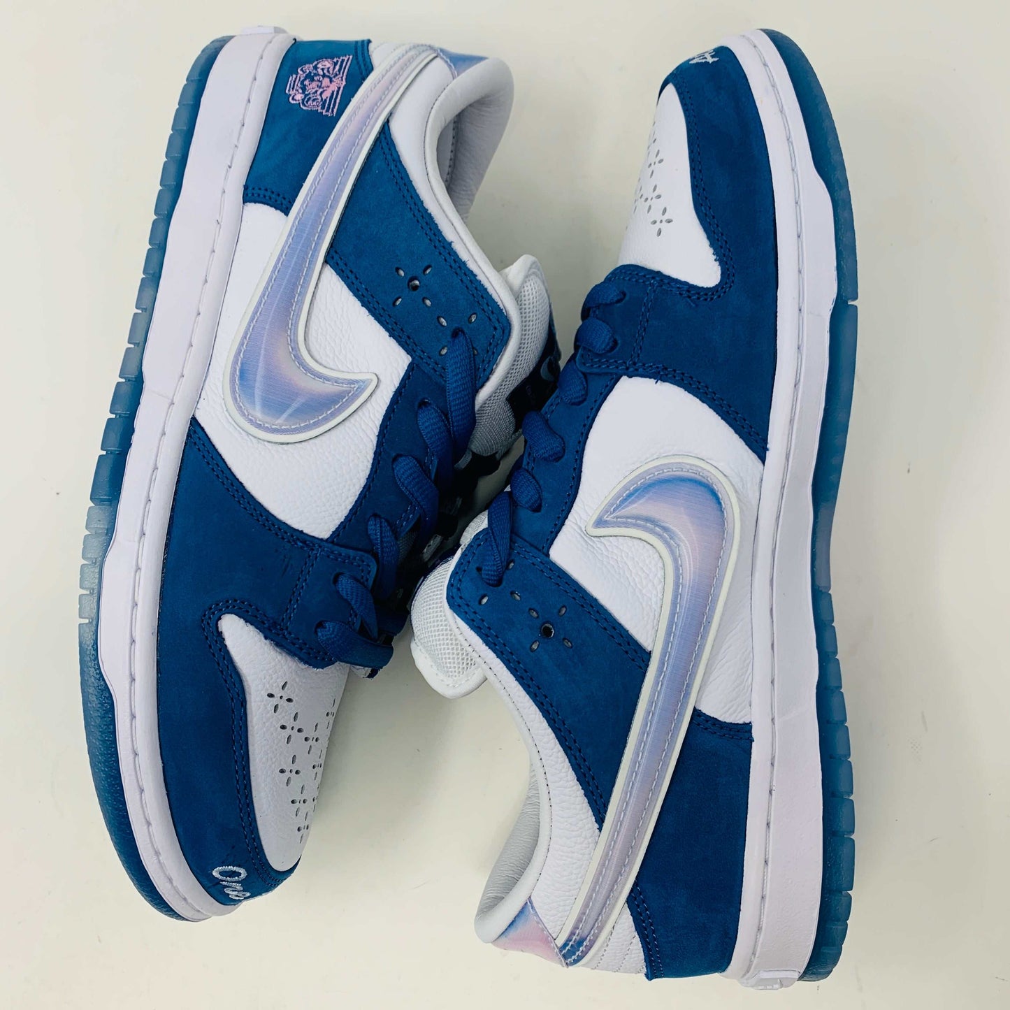 The Nike SB Dunk Low Born X Raised One Block At A Time sneaker, in blue and white with a metallic swoosh and embroidered heel details, sits on a Nike shoebox. It includes three pairs of shoelaces—two white and one pink—in front of the 2023 edition box.