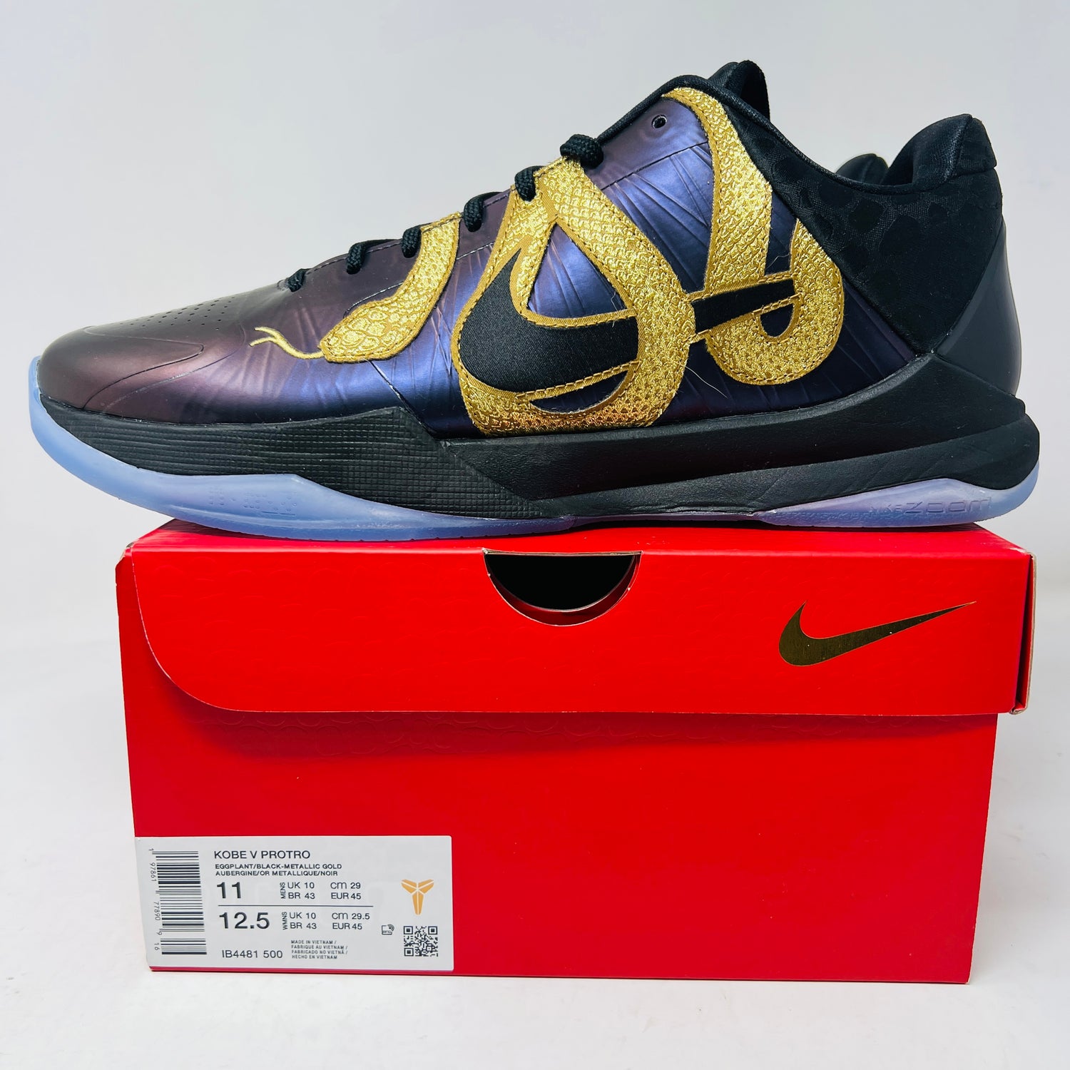 A pristine Nike Kobe 5 Protro Year of the Mamba Eggplant sneaker, featuring a black base with intricate gold patterns and a translucent sole, sits atop its red Nike shoebox. The box label highlights size and style, making it attractive for accessories enthusiasts.