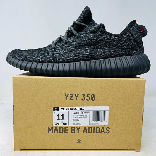 A genuine adidas Yeezy Boost 350 Pirate Black (2023) sneaker features a textured design with red stitching on the heel tab. It sits on a brown YZY 350 and MADE BY ADIDAS shoebox, displaying the shoe size and barcode.