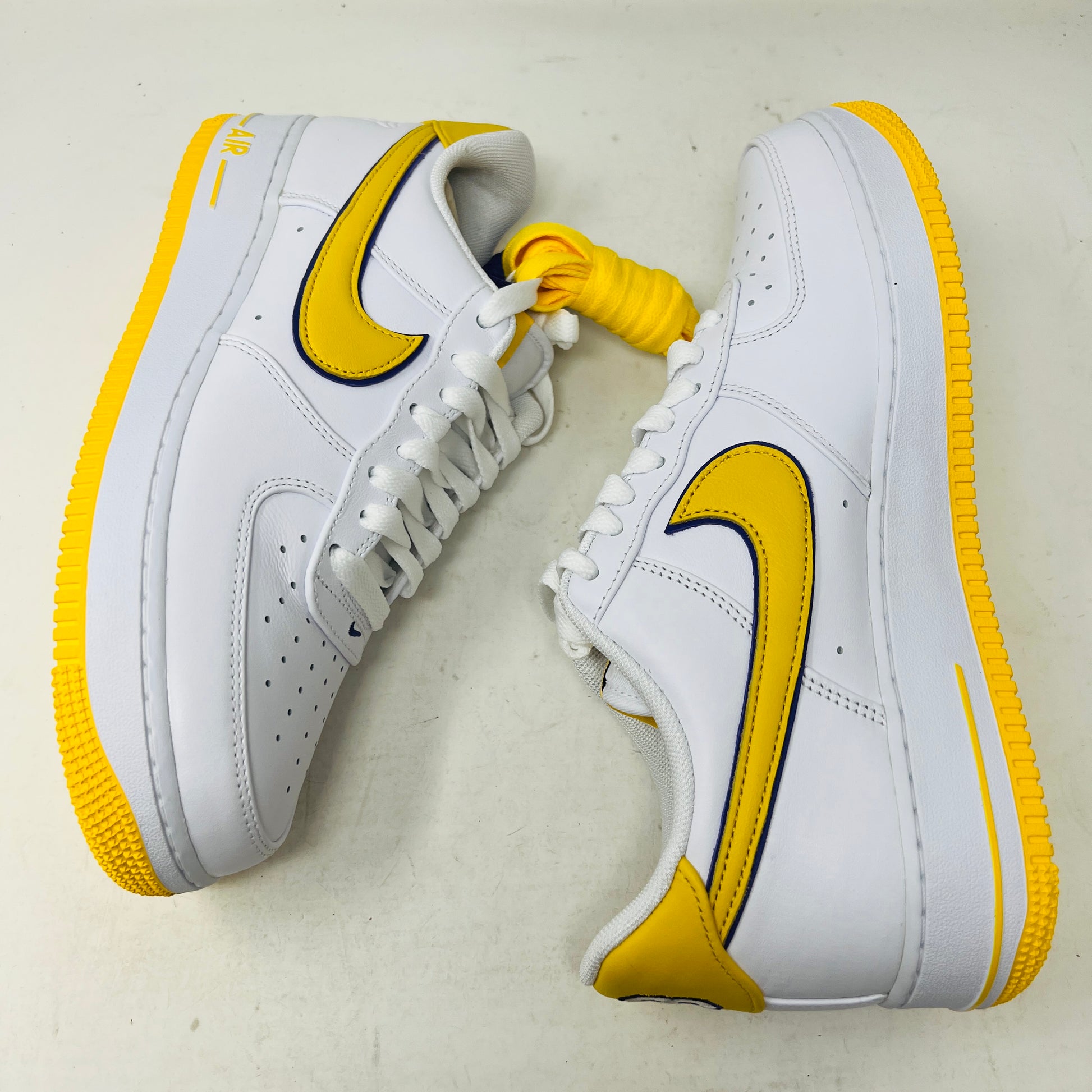 The Nike Air Force 1 Low Retro QS Kobe Bryant Lakers Home sneakers, featuring white with yellow swooshes and soles, are displayed against a plain background with an outer side and top view.