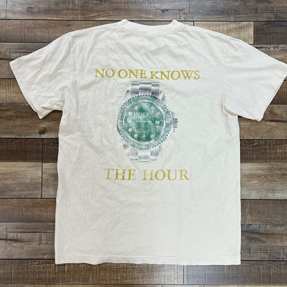 A cream-colored Holy Ground Holex Tee Regular with HOLY GROUND in green on the chest is displayed on a rustic wooden surface.