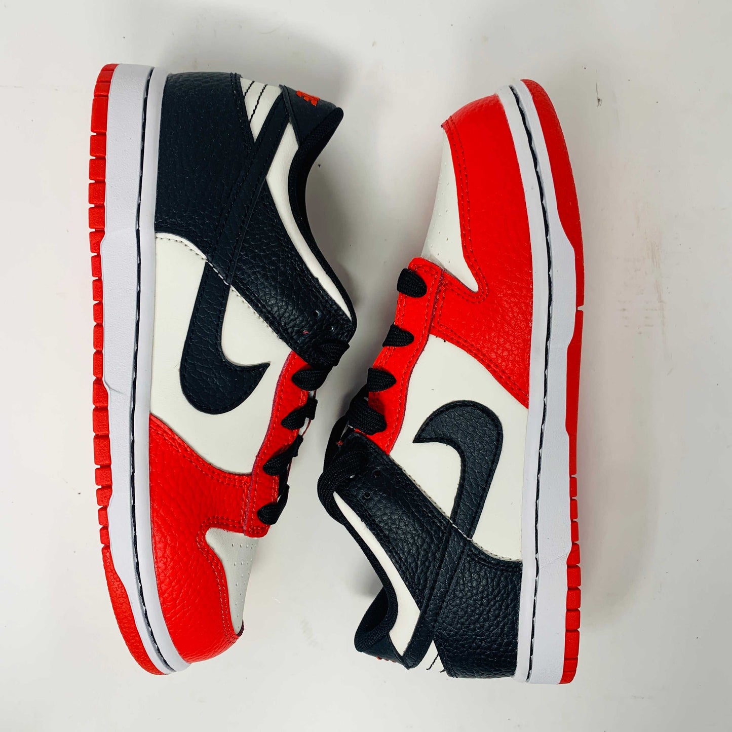 The Nike Dunk Low EMB Chicago GS, a genuine pair of red, black, and white sneakers featuring a sleek black swoosh and matching laces, elegantly sits atop a new red box adorned with bold white Nike branding.