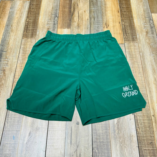 The Holy Ground Athletic Shorts (S24) feature a green design with Holy Ground printed in white on the lower left front, displayed on a wooden floor of brown and tan planks.