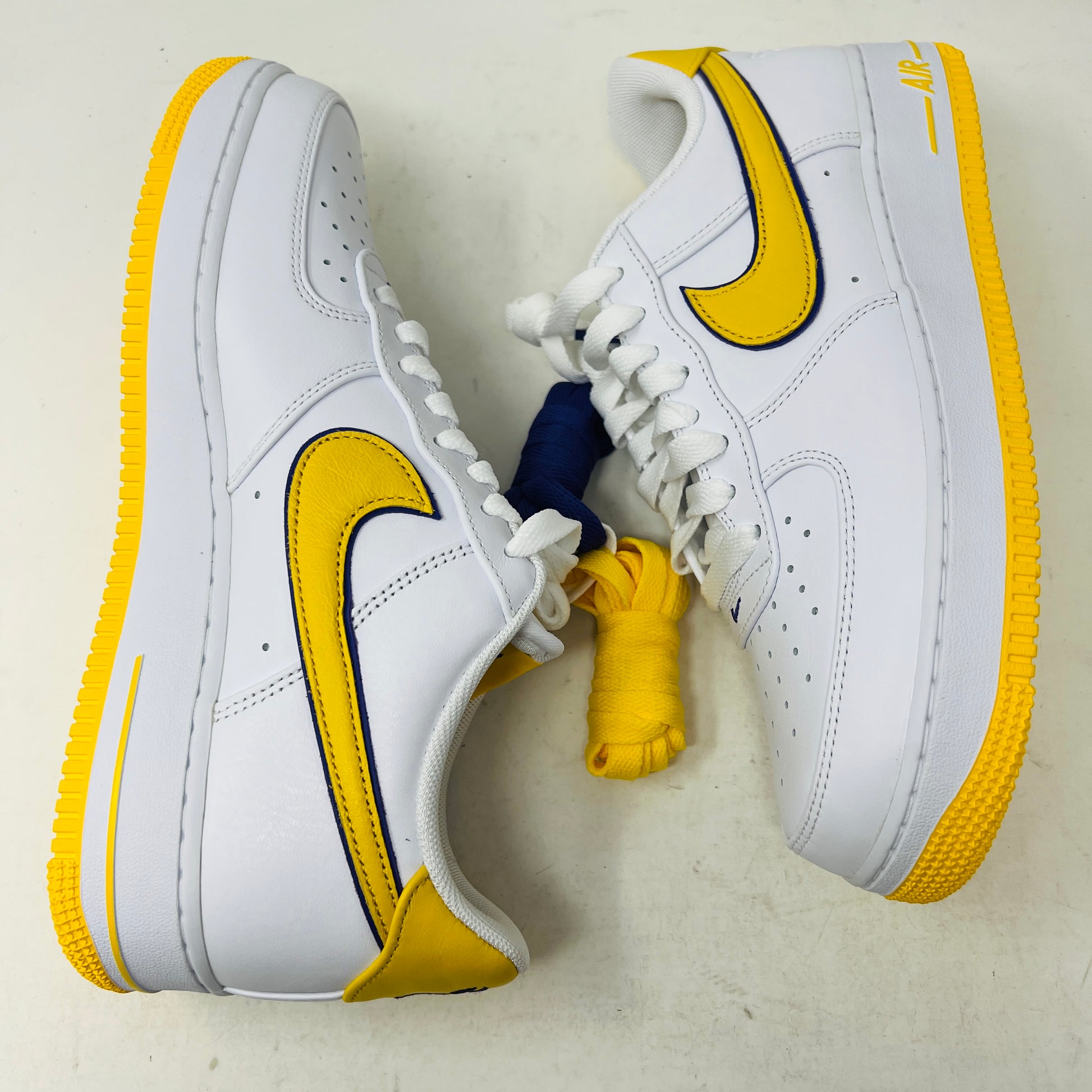 The Nike Air Force 1 Low Retro QS Kobe Bryant Lakers Home features a bright yellow swoosh and yellow detailing on a white background. It includes two extra pairs of laces in blue and yellow.