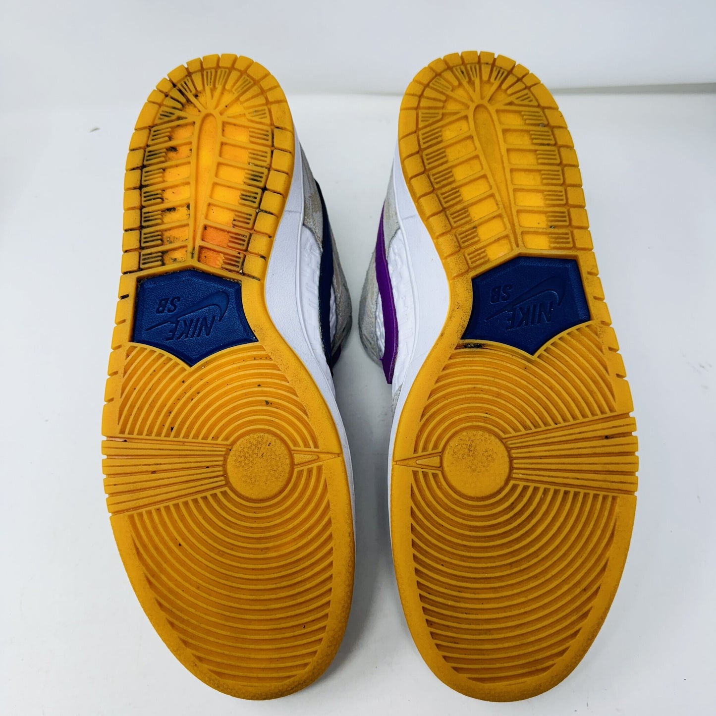 The image showcases the soles of authentic Nike SB Dunk Low Rayssa Leal sneakers. The predominantly yellow soles with blue accents near the heels and circular patterns on the toes show wear. However, the clean uppers suggest well-maintained size 12M sneakers in great condition.