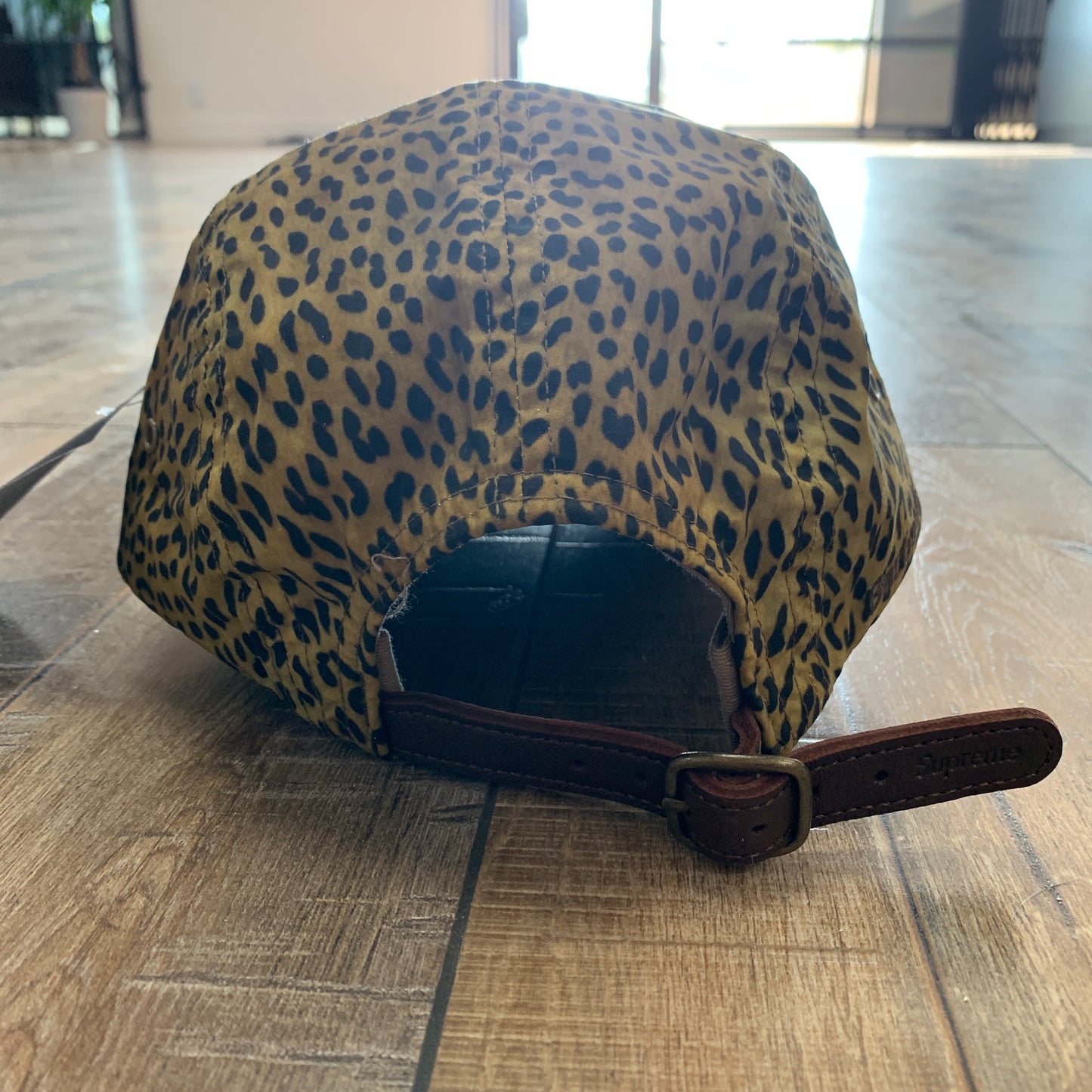 The Supreme Barbour Waxed Cotton Camp Cap Leopard with an adjustable strap rests on a wooden floor near a pair of brand new authentic shoes, as sunlight streams in from a large open doorway in the background.