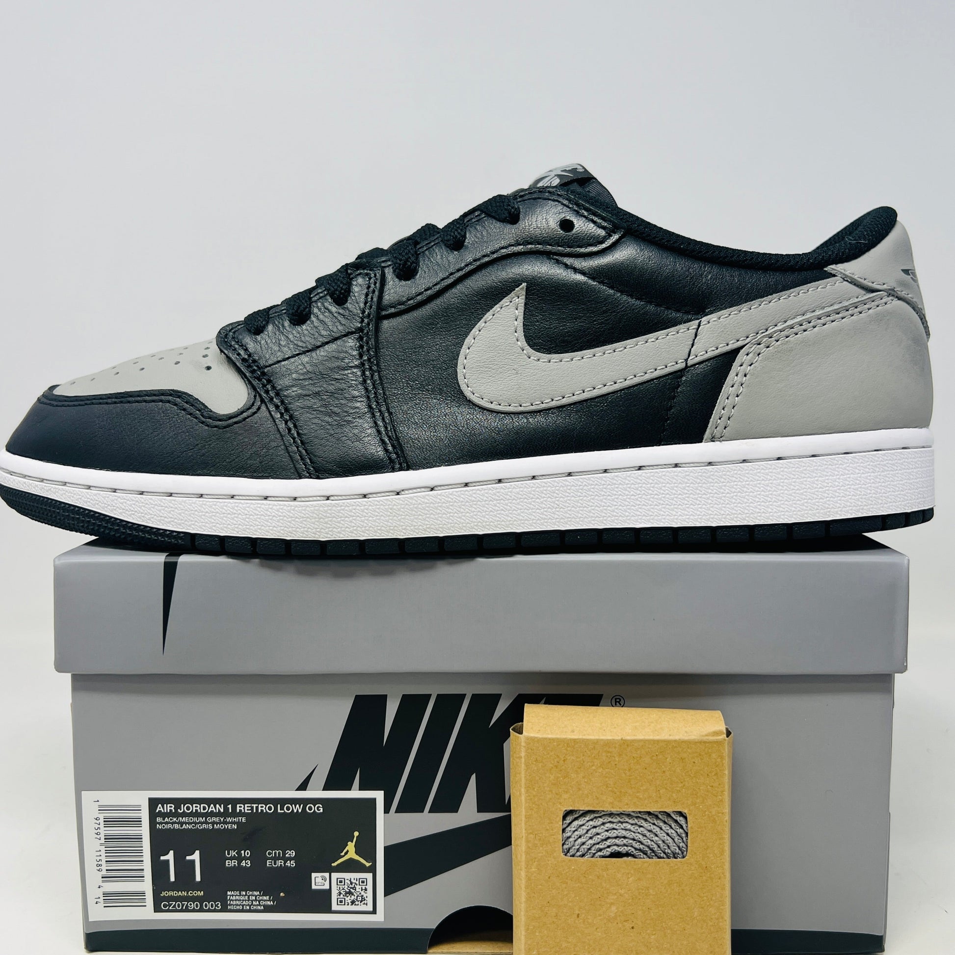 The Jordan 1 Retro Low OG Shadow (2024) sneakers in black and grey are displayed on a grey Nike box, showcasing clean uppers and matching laces. A small brown box complements the scene, with the visible size label enhancing authenticity.