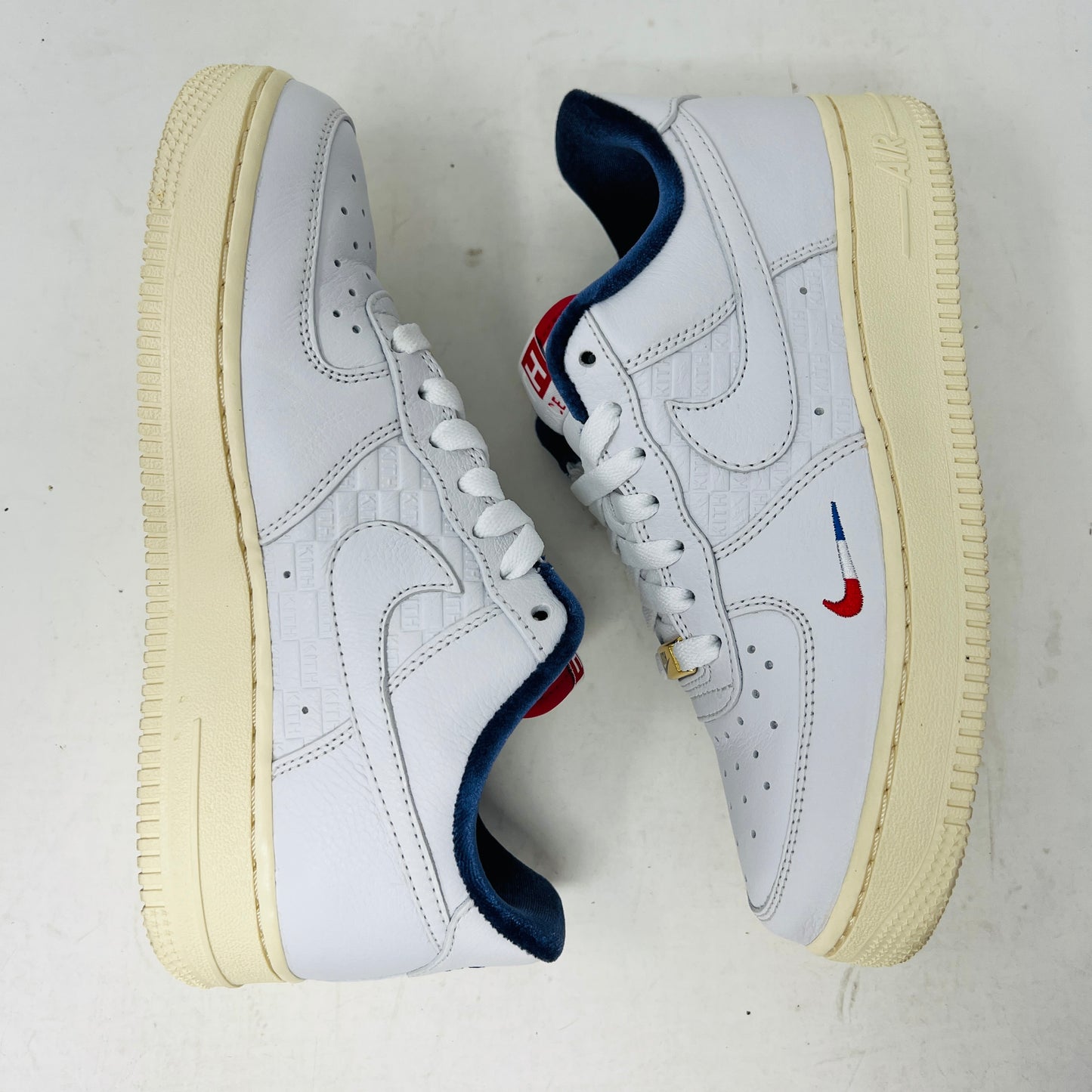 The Nike Air Force 1 Low Kith Paris sneakers feature embossed patterns and blue and red detailing on a white body with off-white soles, set against a white background.