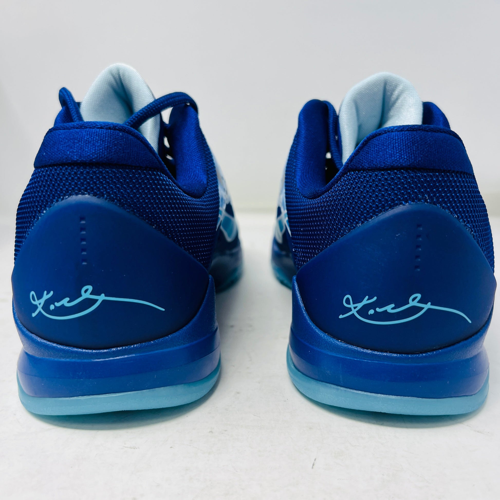 The Nike Kobe 5 X-Ray (GS) athletic shoes showcase a back view of their blue design with light blue soles, each featuring a signature on the heel. They are presented on a white surface against a light background.