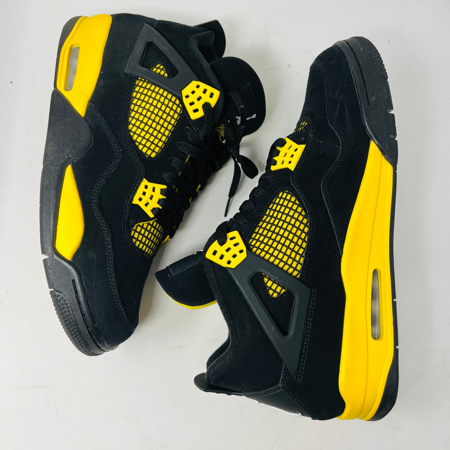 The Jordan 4 Retro Thunder (2023) features black uppers with yellow accents, a white sole, and visible air cushioning. It rests on a black and gray speckled shoebox in good condition and includes the distinctive wing design.