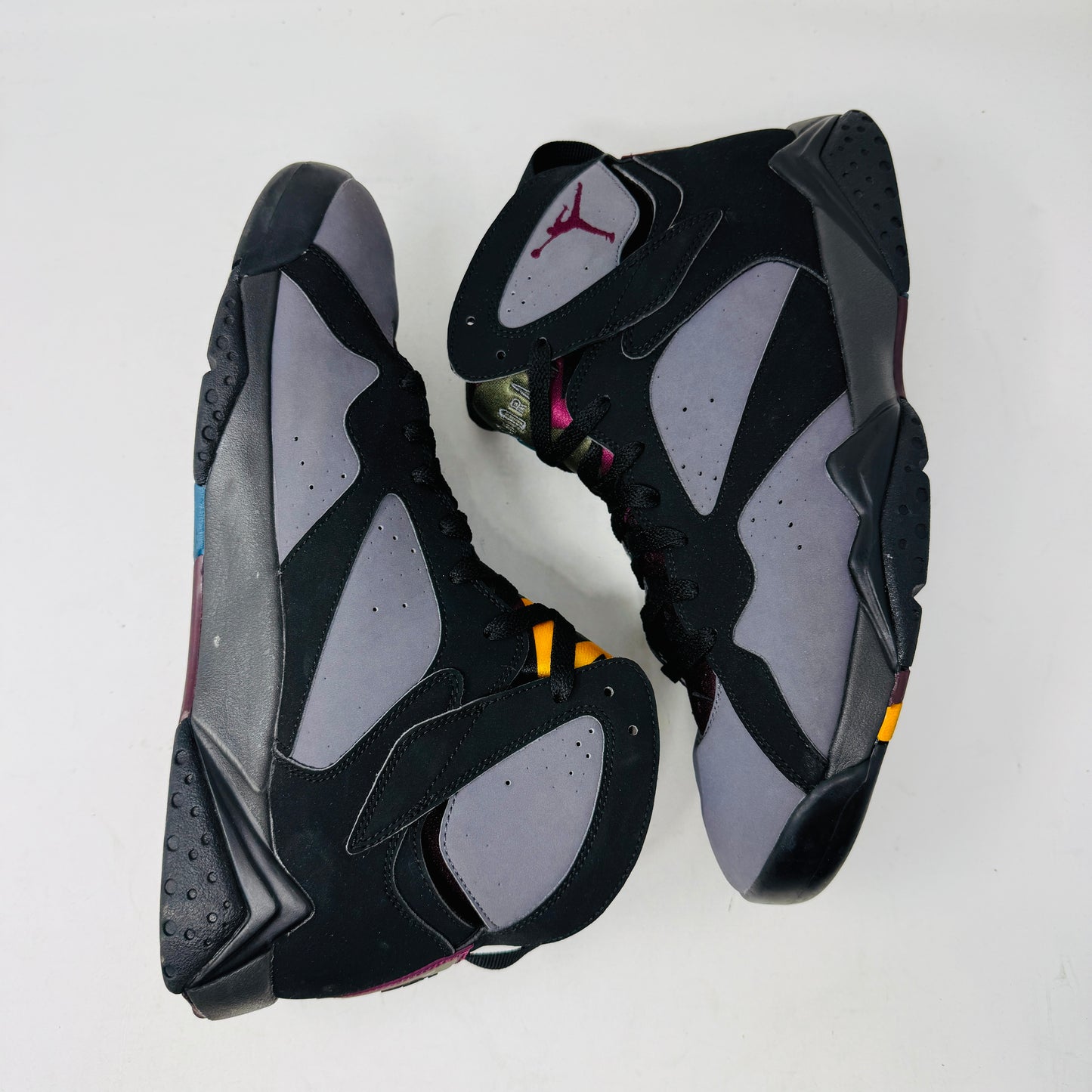 The Jordan 7 Retro Bordeaux (2015) sneakers, in black and gray with maroon accents and the Jumpman logo, feature angular midsoles, dark laces, and sleek uppers. They evoke a 2015 vibe and are photographed side by side on a white background.