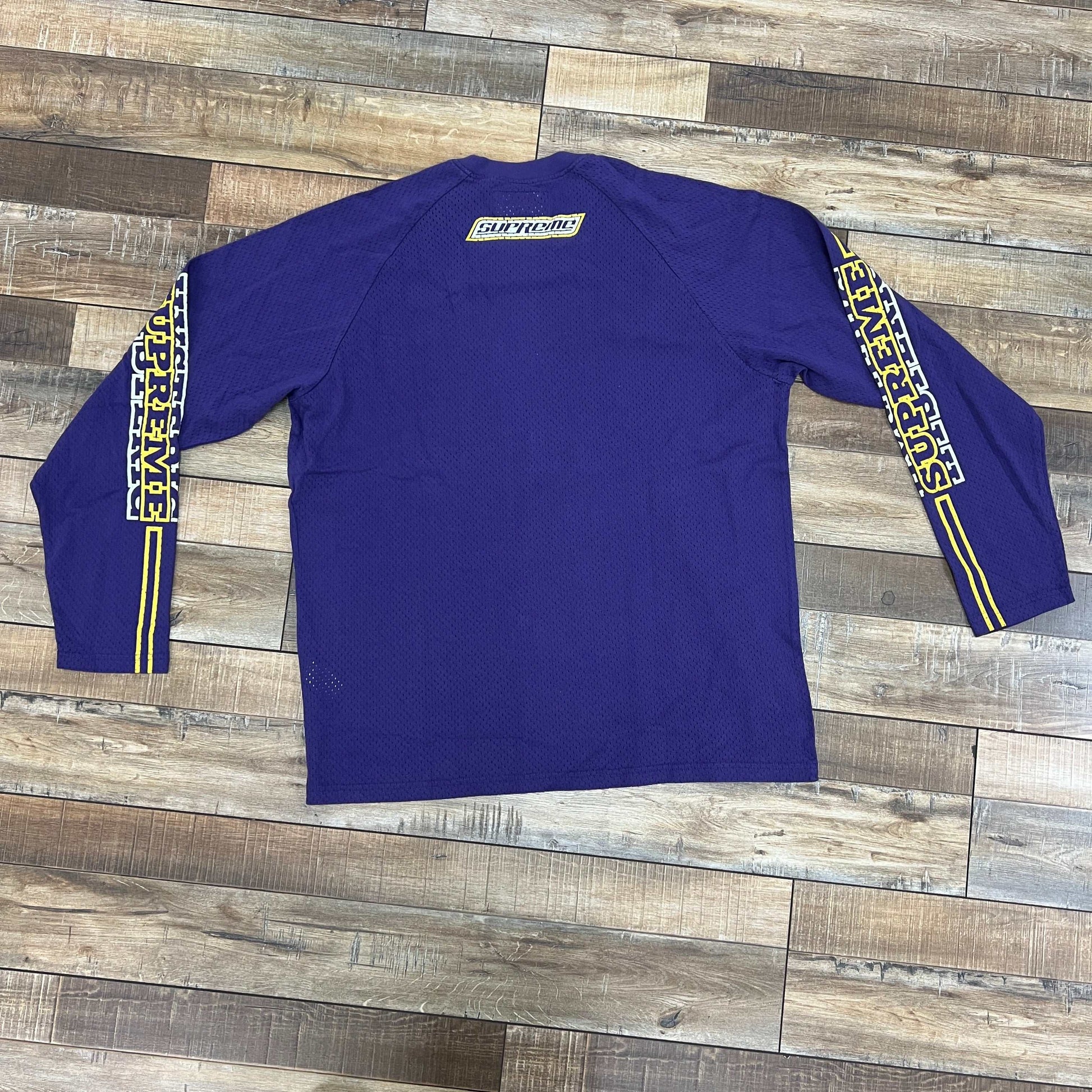 The Supreme Hysteric Glamour Mesh L/S Top in Purple is a brand new, authentic long-sleeve shirt with yellow and white logos on the back and sleeves, displayed flat on wooden flooring to highlight its vibrant design.