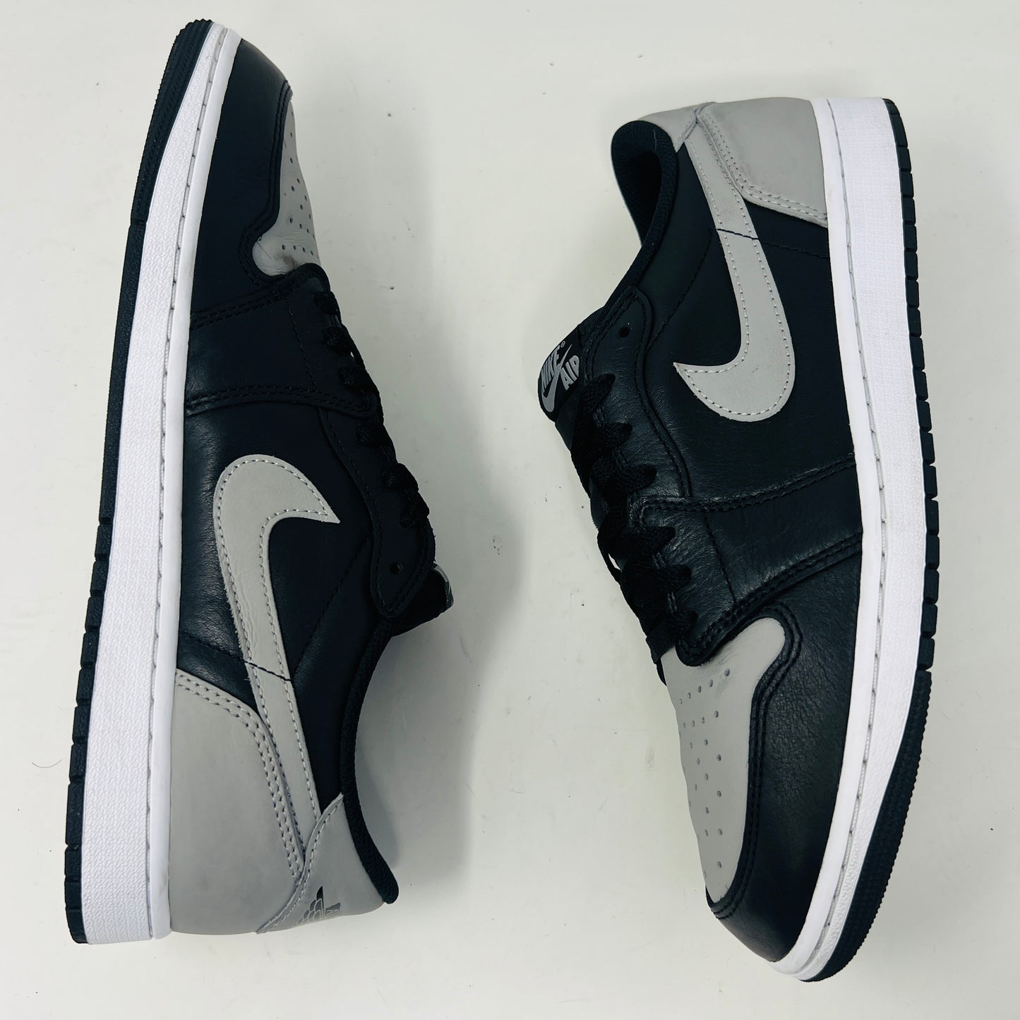 The Jordan 1 Retro Low OG Shadow (2024) sneakers in black and grey are displayed on a grey Nike box, showcasing clean uppers and matching laces. A small brown box complements the scene, with the visible size label enhancing authenticity.